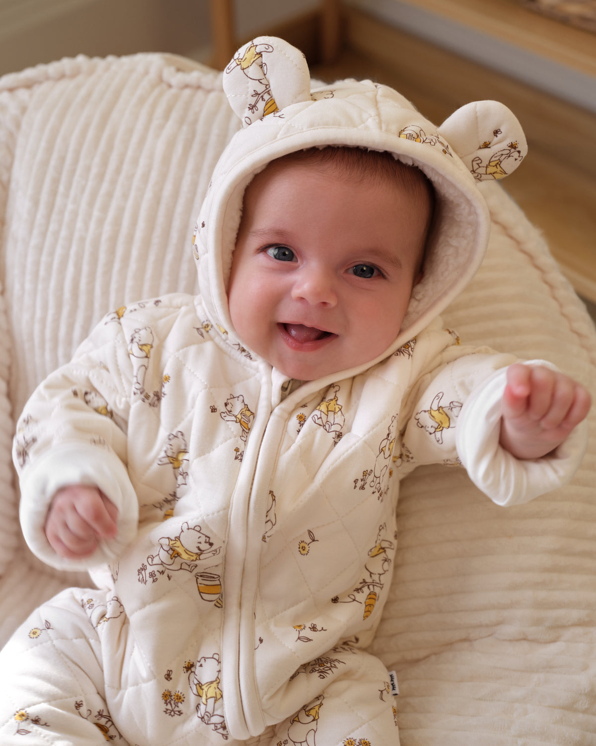 A baby wearing a cream-colored hooded snowsuit with a honey bee pattern, sitting on a ribbed cream cushion.