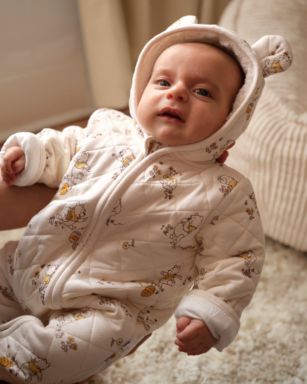 A baby wearing a cream-colored quilted hooded snowsuit with a honeybee and bee print, sitting on a soft beige rug.