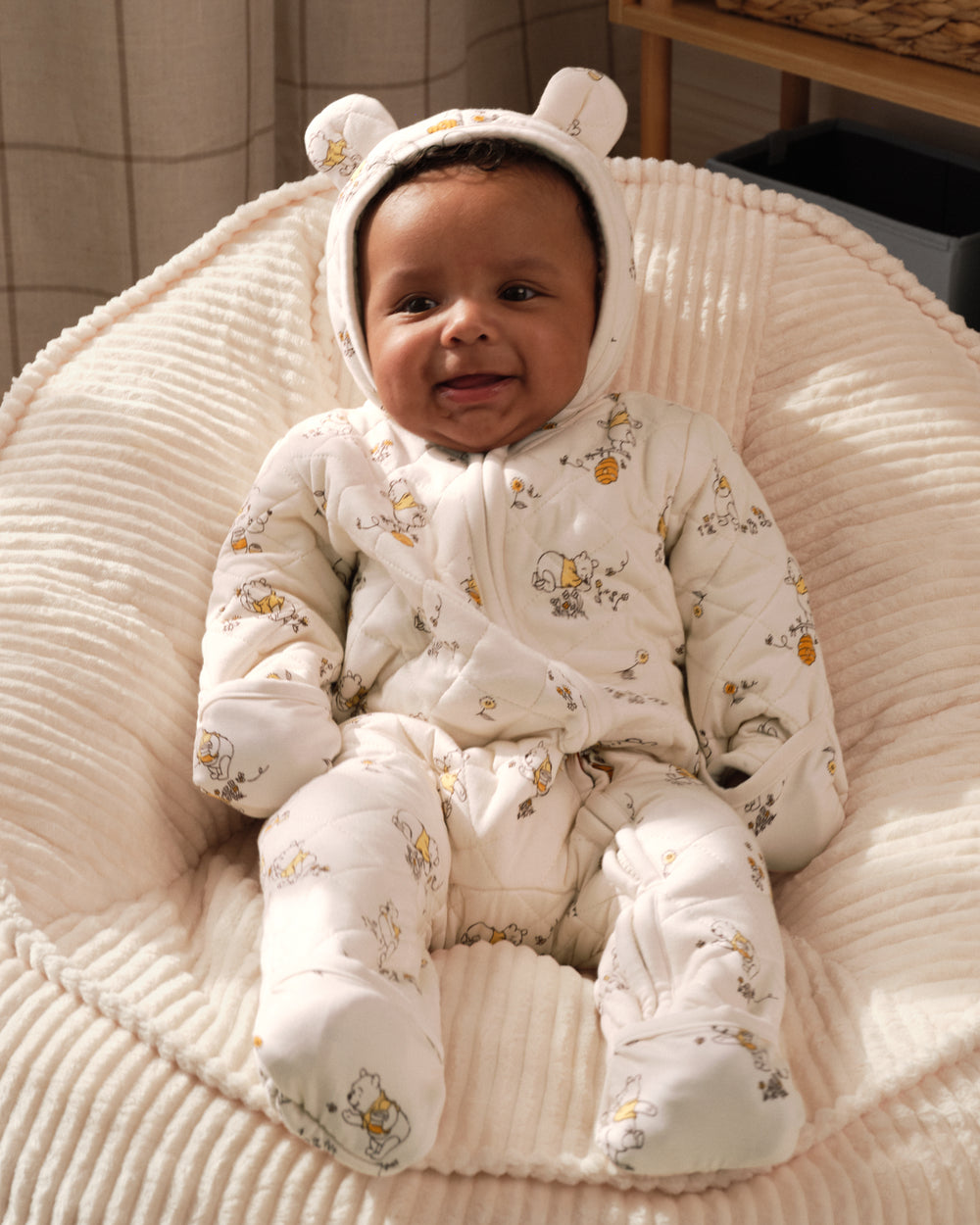 A baby wearing a white quilted hooded snowsuit with a small animal print, sitting in a cream ribbed fabric bassinet or chair.