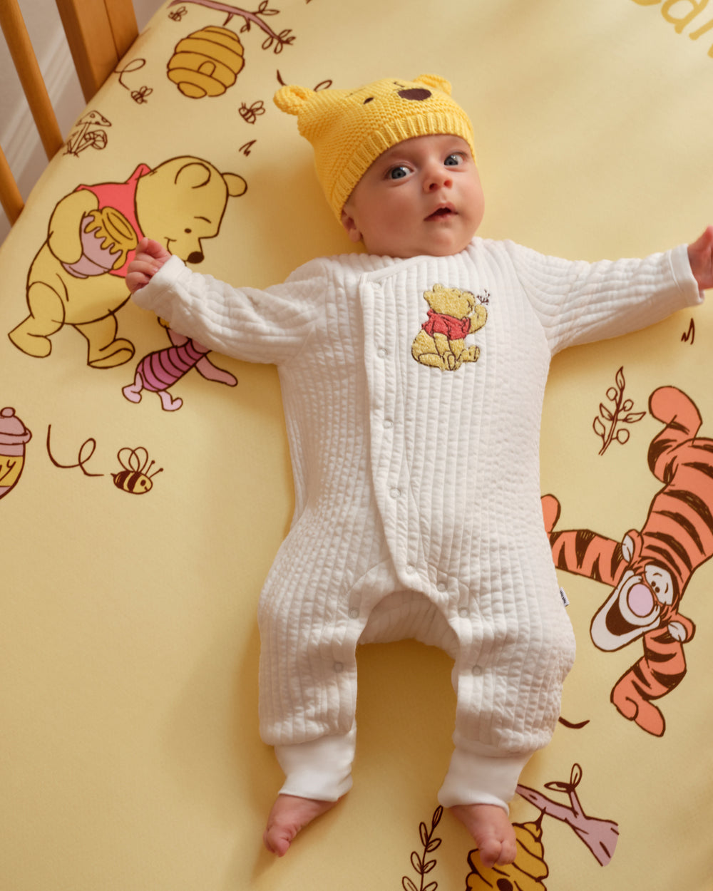 A baby lying on a Winnie the Pooh themed yellow blanket, wearing a white textured long-sleeve footless sleeper with a Winnie the Pooh chest print and a yellow knit Pooh hat.