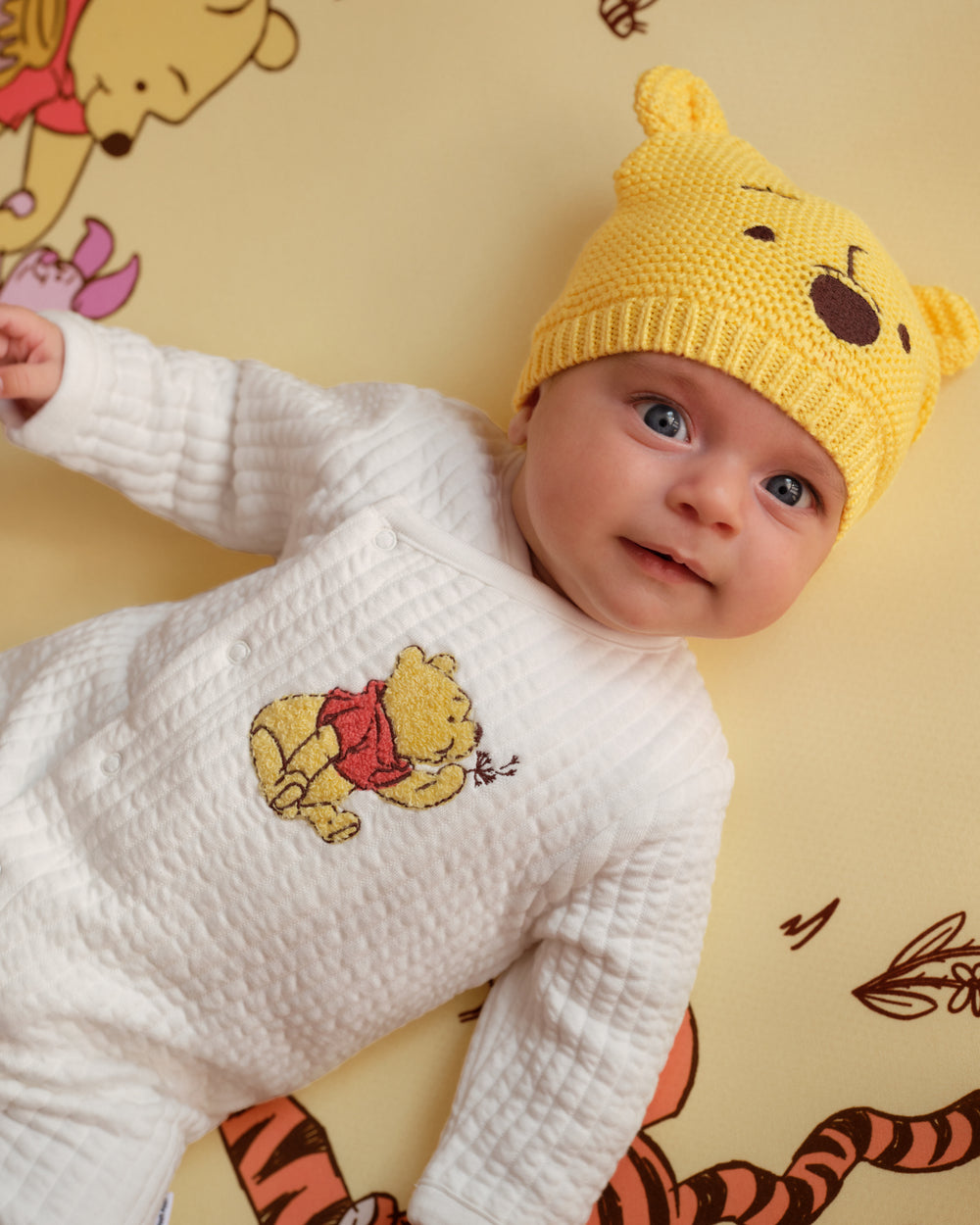 A baby lying on a yellow Winnie the Pooh themed blanket, wearing a white quilted long-sleeve bodysuit with a Pooh embroidery and a yellow knit Pooh hat.
