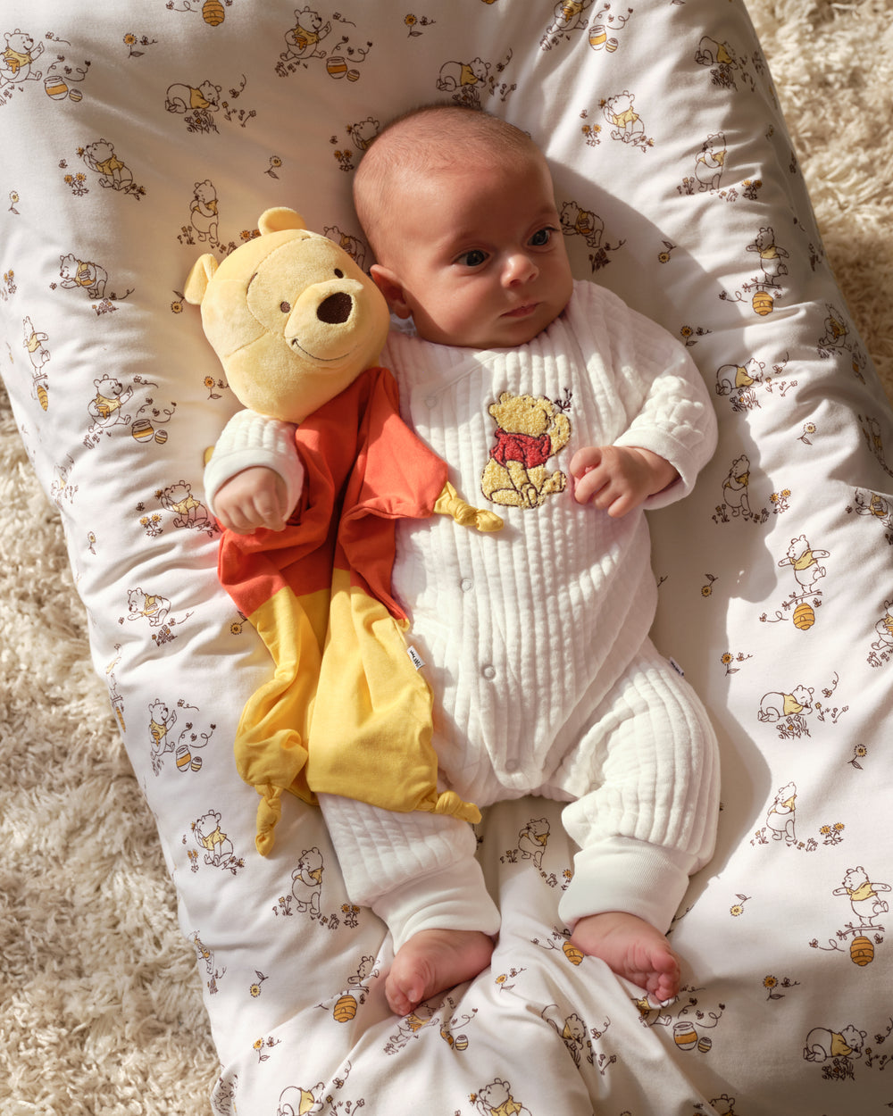A baby lying on a white patterned baby lounger, wearing a white ribbed onesie with a Winnie the Pooh graphic, and holding a plush Winnie the Pooh toy.