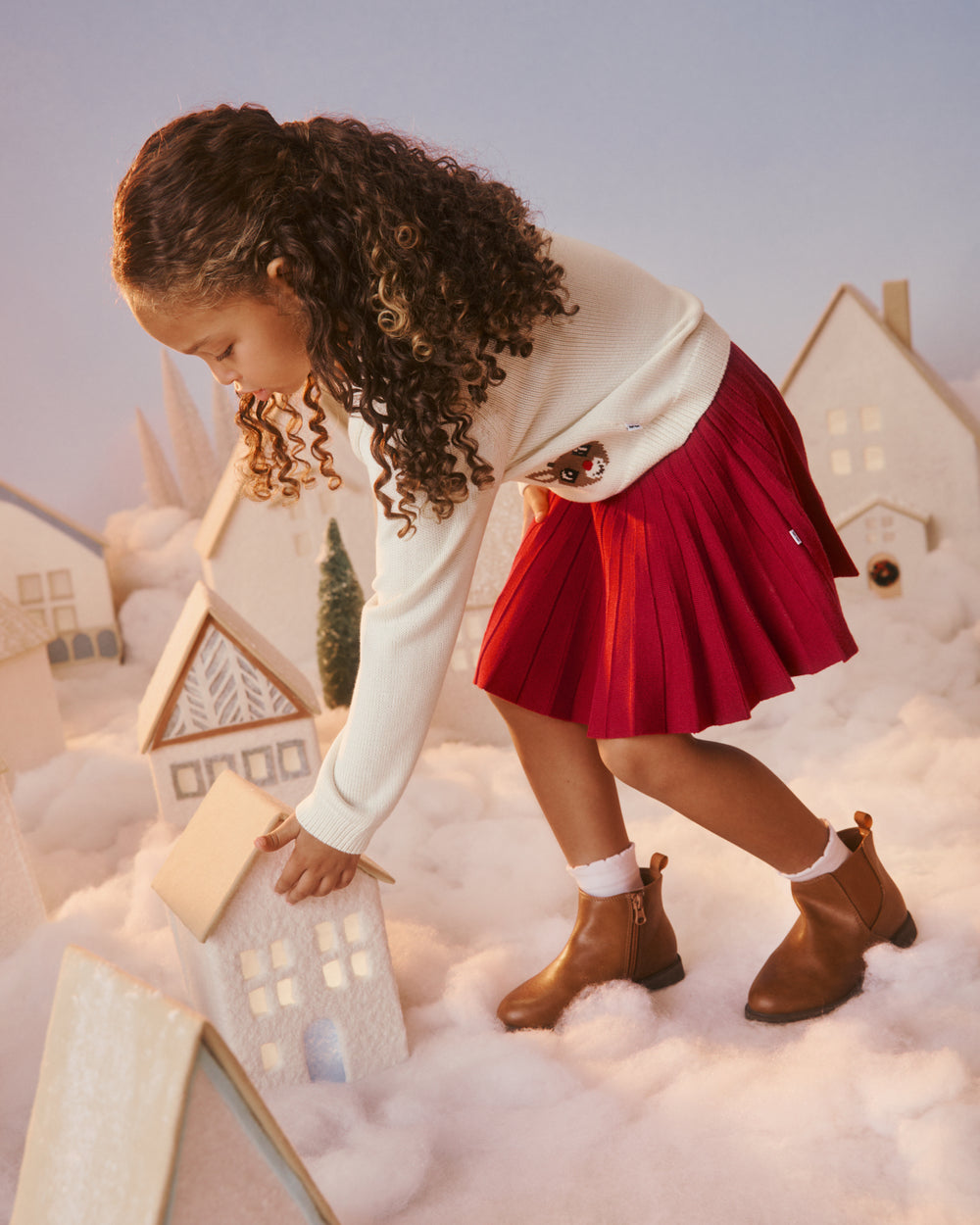 A young girl shown from a side profile, wearing a white long-sleeve top, a red pleated skirt, white socks, and brown ankle boots. 