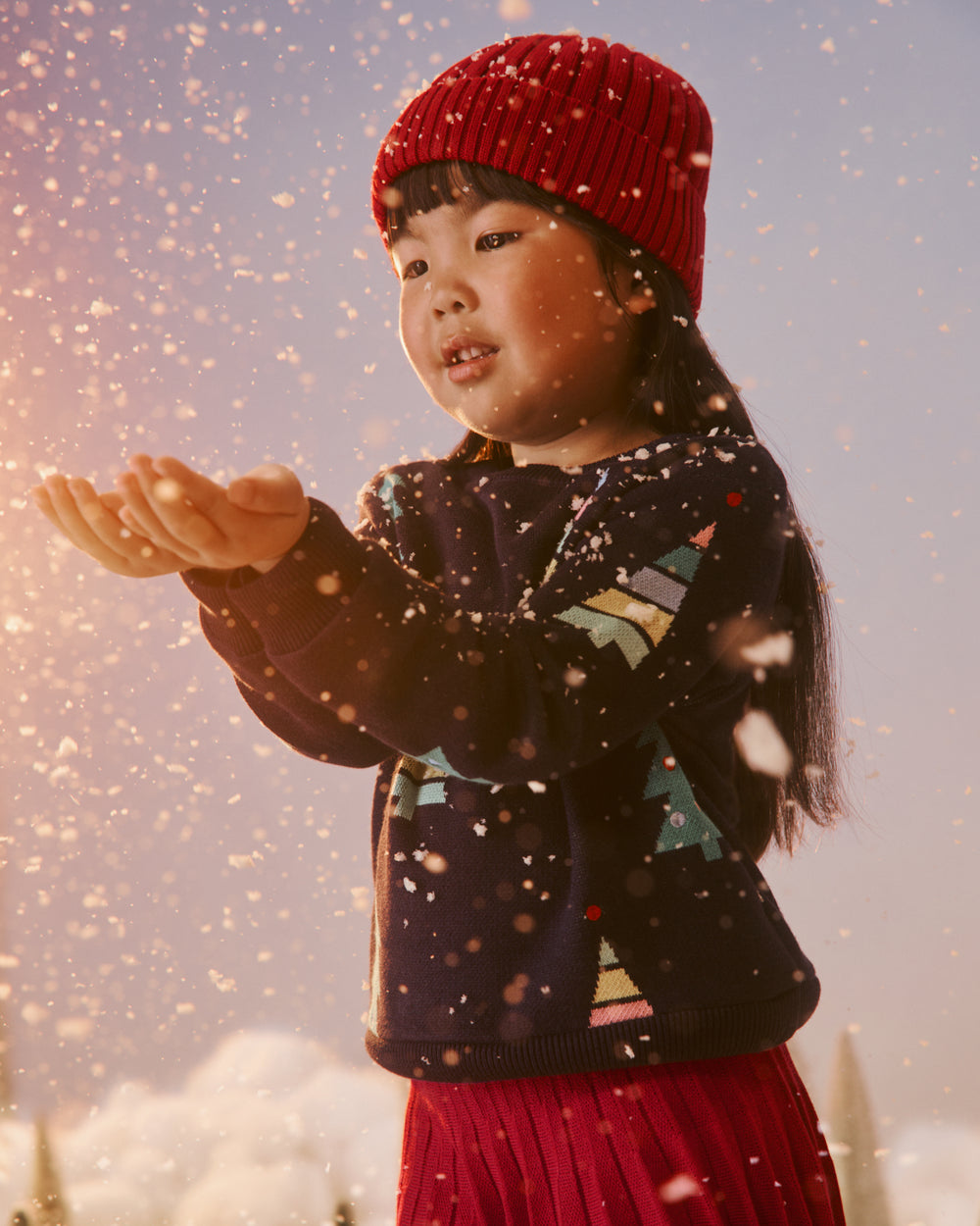 A young girl wearing a red knit beanie, a dark navy sweater with colorful geometric patterns, and a red pleated skirt stands outdoors in falling snow, holding her hands out to catch the flakes.