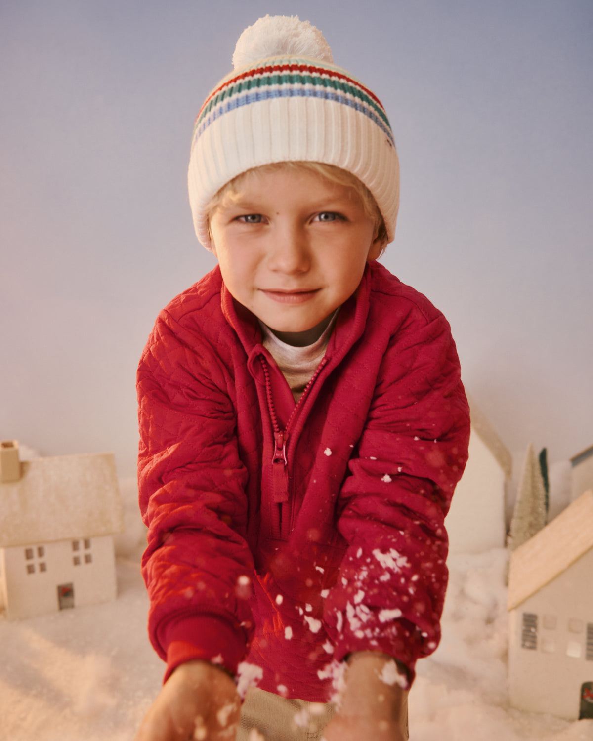 A young child wearing a white knit beanie with colorful stripes and a red winter jacket, reaching toward snow in a snowy outdoor setting with miniature houses in the background.