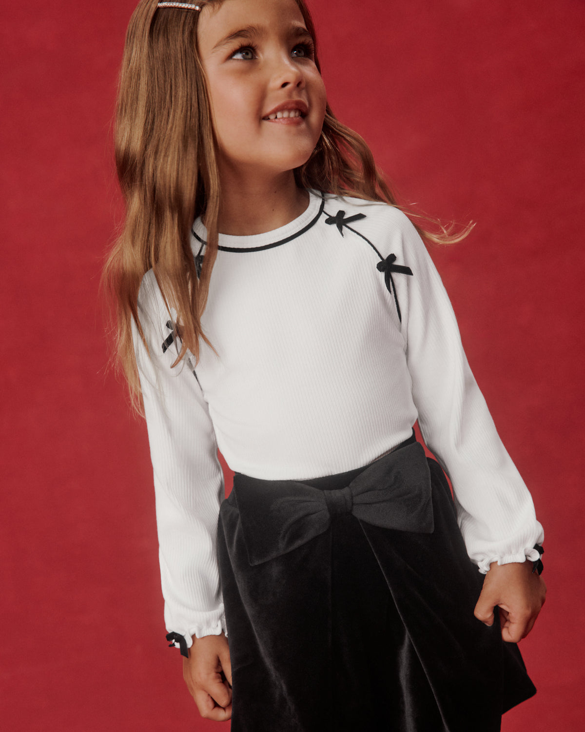 A young girl with long light brown hair wearing a white long-sleeve top with black embroidered dragonflies and a black skirt with a large bow, posing against a textured red background.