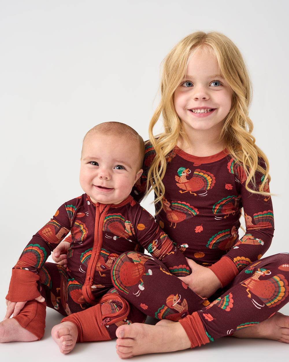 Two children sitting side by side wearing matching long-sleeve pajamas with a brown background and a colorful turkey print. The pajamas have orange zippers and cuffs, and the children are barefoot against a plain light gray studio background.