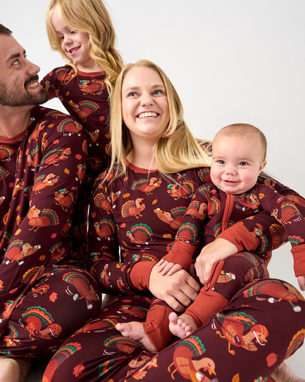A family of four wearing matching long-sleeve pajamas with a brown background and a colorful autumn pattern featuring squirrels, turkeys, and leaves, posed together against a plain light gray background.