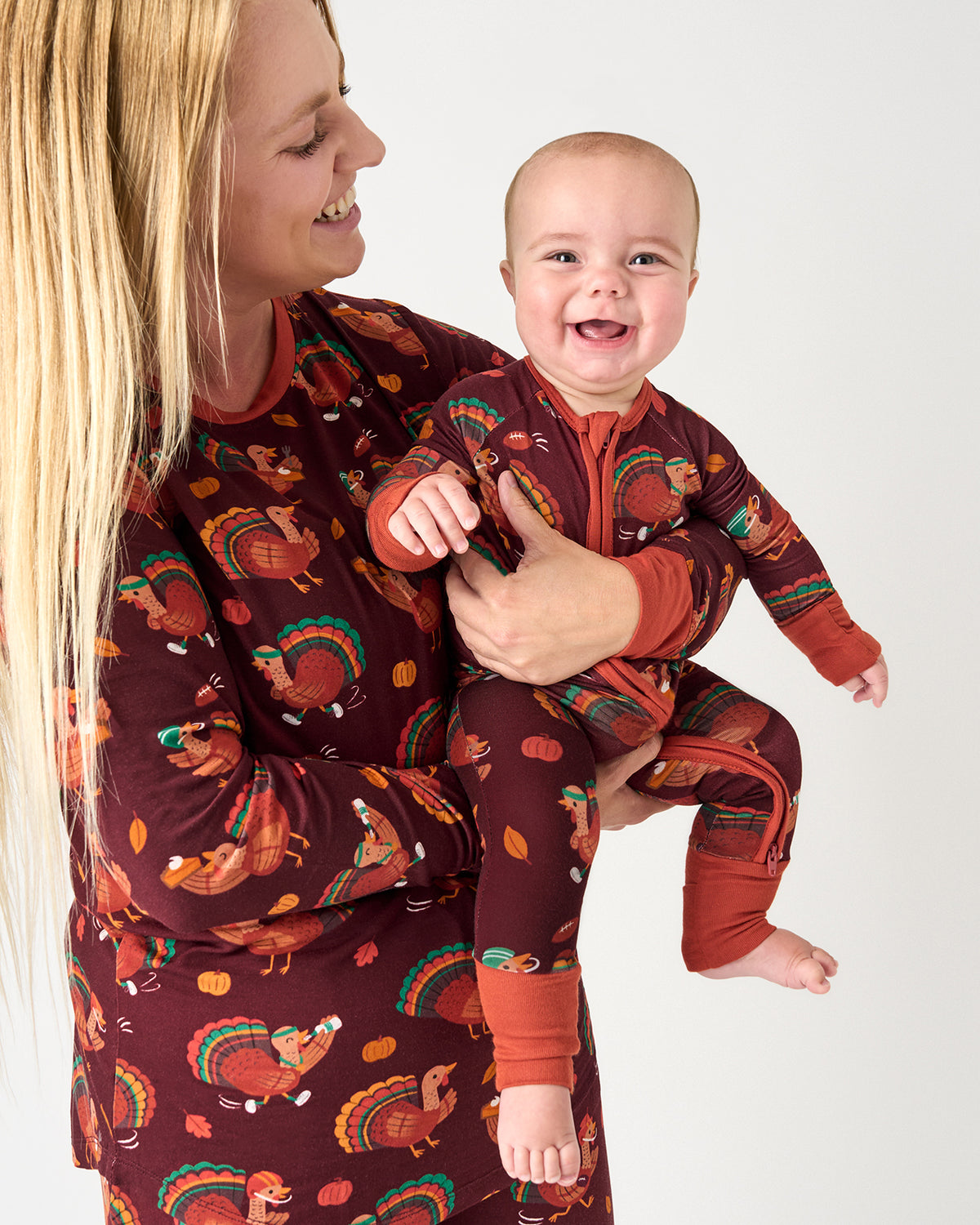 A smiling woman holding a baby wearing matching brown and rust-colored pajamas with a turkey and autumn leaf print, against a plain light gray background.