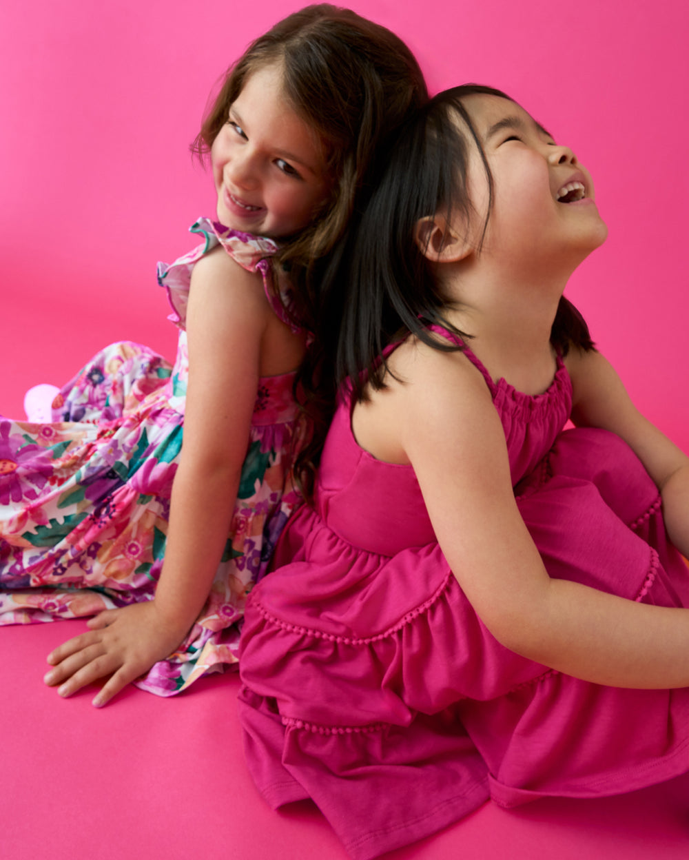 Two sitting girls wearing dresses from the Summer Capsule on a pink backdrop 
