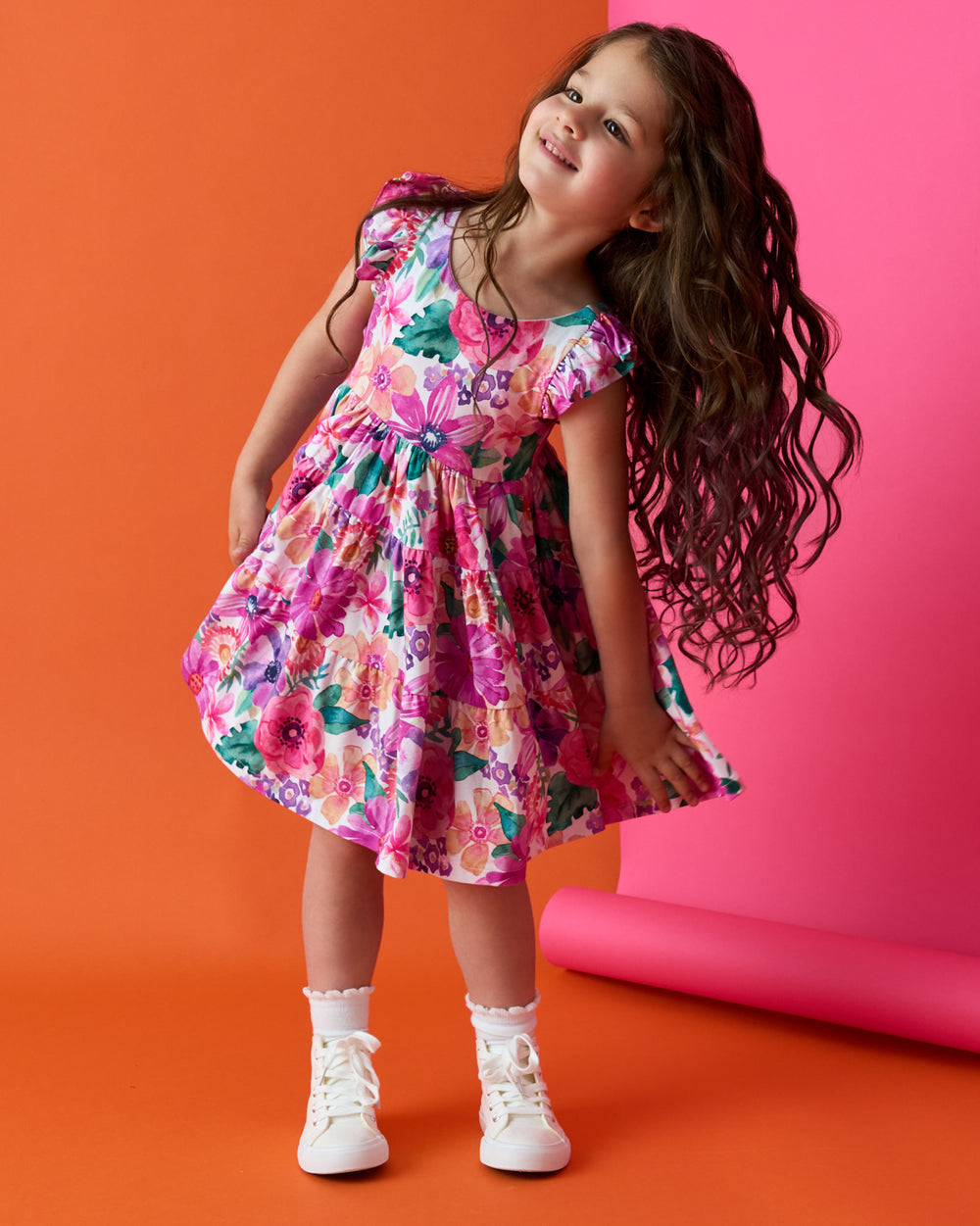 Girl wearing the Hibiscus Blooms Tiered Flutter Dress on an orange and pink back drop