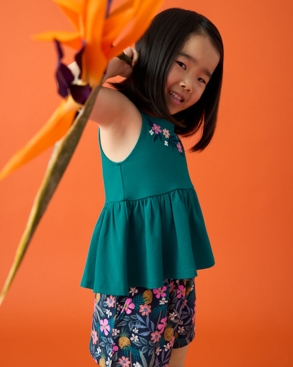 Girl wearing the Tropical Garden Smocked Shorts and green tank top on a orange backdrop 