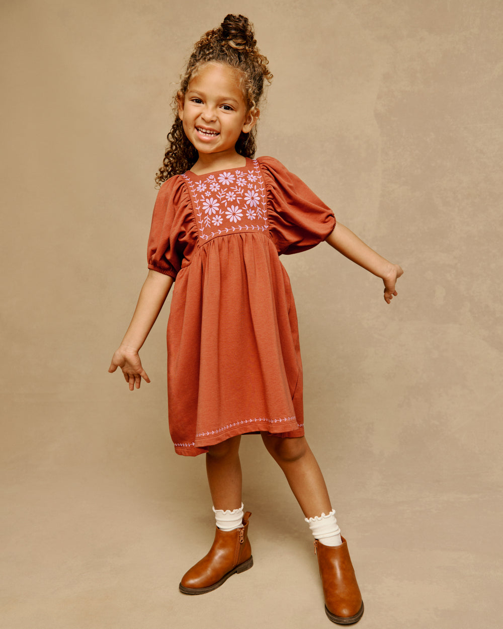 A young girl is modeling a rust-colored short-sleeve dress with floral embroidery on the chest, styled with white socks and brown boots against a textured beige studio backdrop.