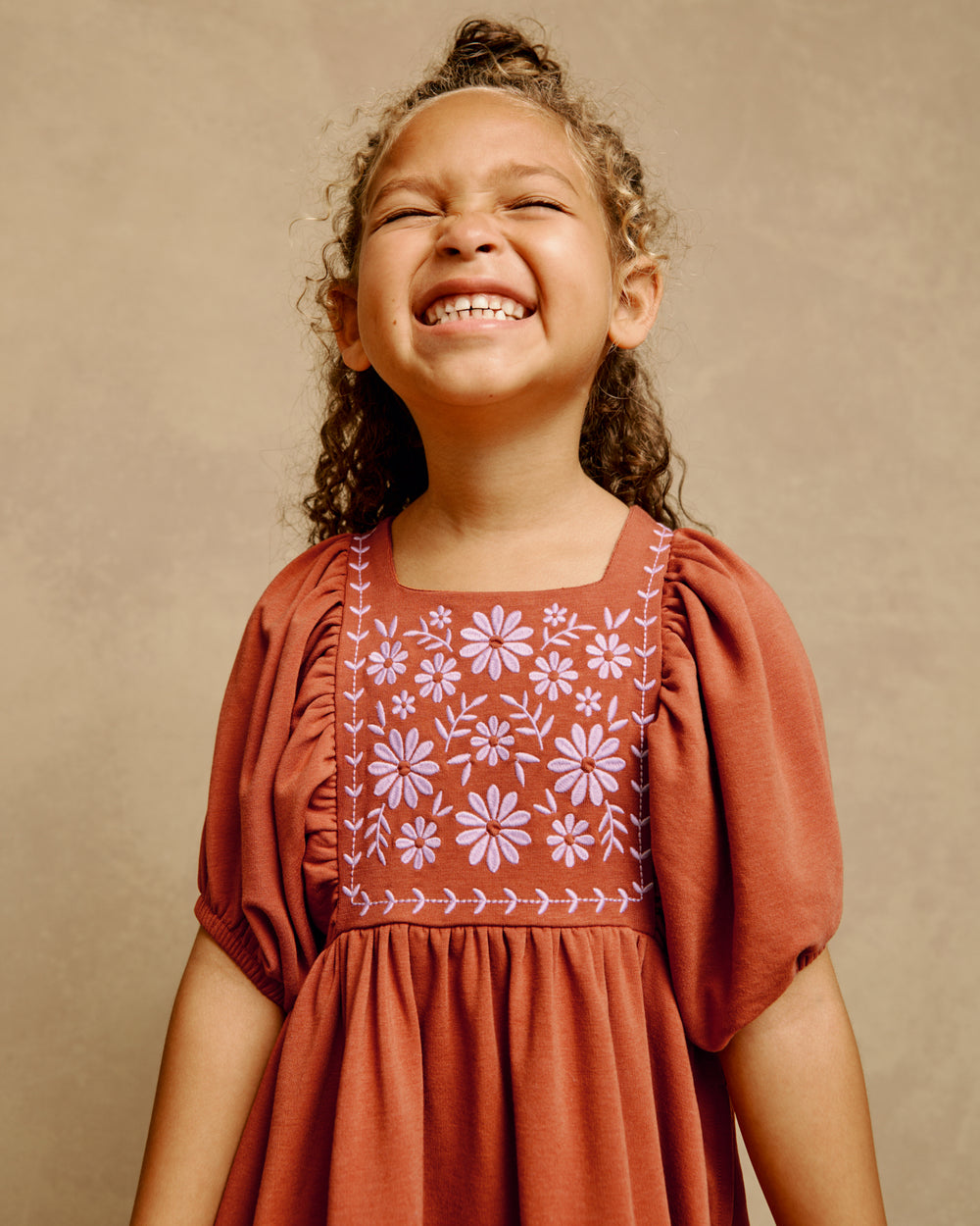 A young girl smiling broadly, wearing a terracotta-colored dress with floral embroidery on the chest, set against a warm beige textured background.
