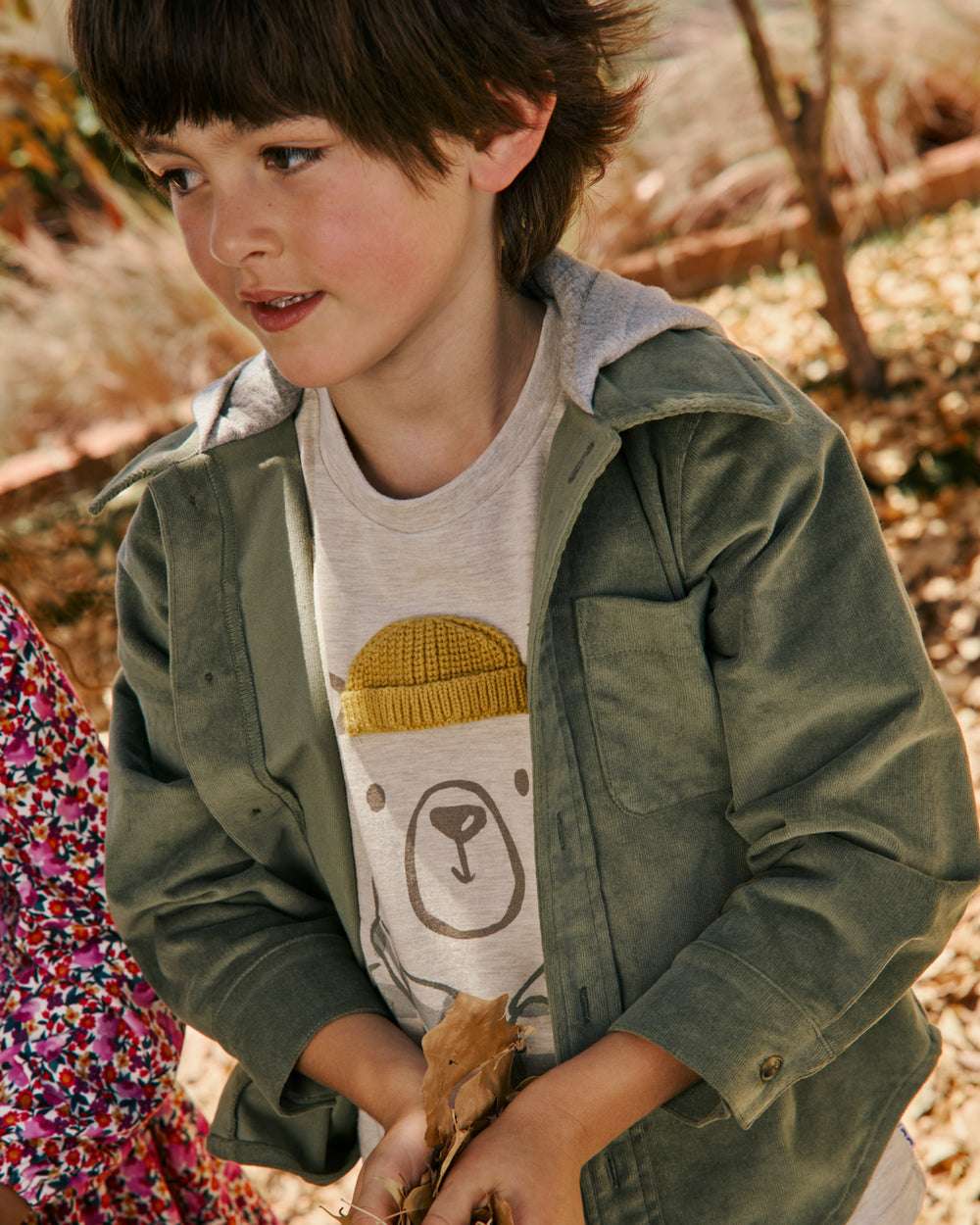 A young boy wearing an olive green hooded jacket over a light gray graphic t-shirt, standing outdoors in a wooded setting.