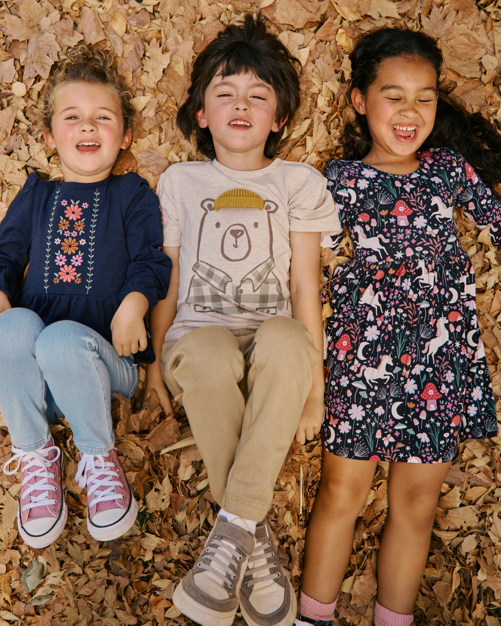 Three children lying on a bed of dry autumn leaves, wearing colorful clothing. The child on the left wears a navy blue embroidered top, light blue jeans, and pink sneakers.