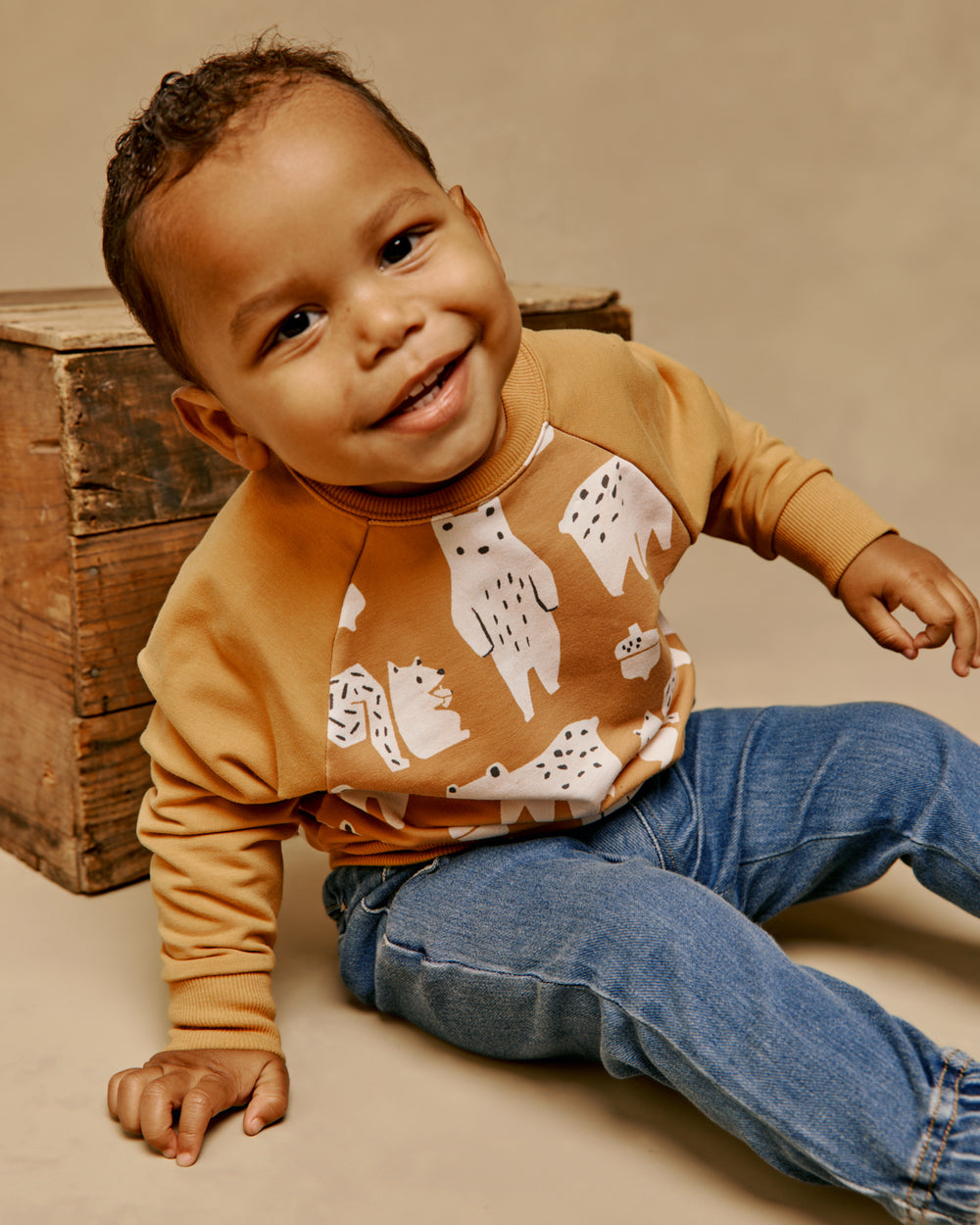 A smiling toddler sitting on a beige floor next to a rustic wooden crate, wearing a mustard yellow and brown long-sleeve raglan sweatshirt with a white forest animal pattern, paired with blue jeans.