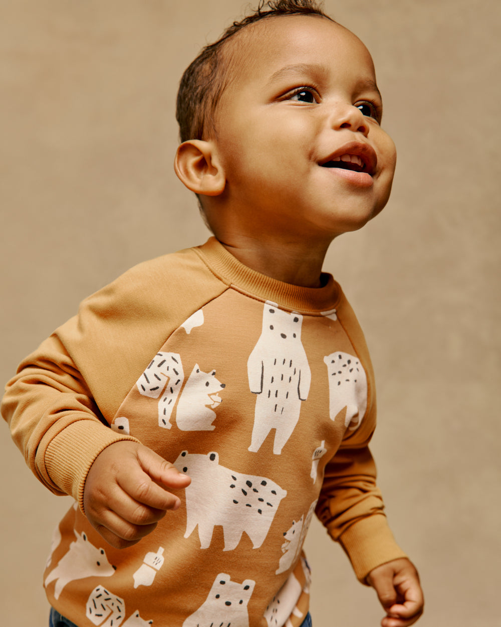 A smiling toddler wearing a tan long-sleeve sweatshirt with a white bear pattern, photographed against a warm beige studio background.