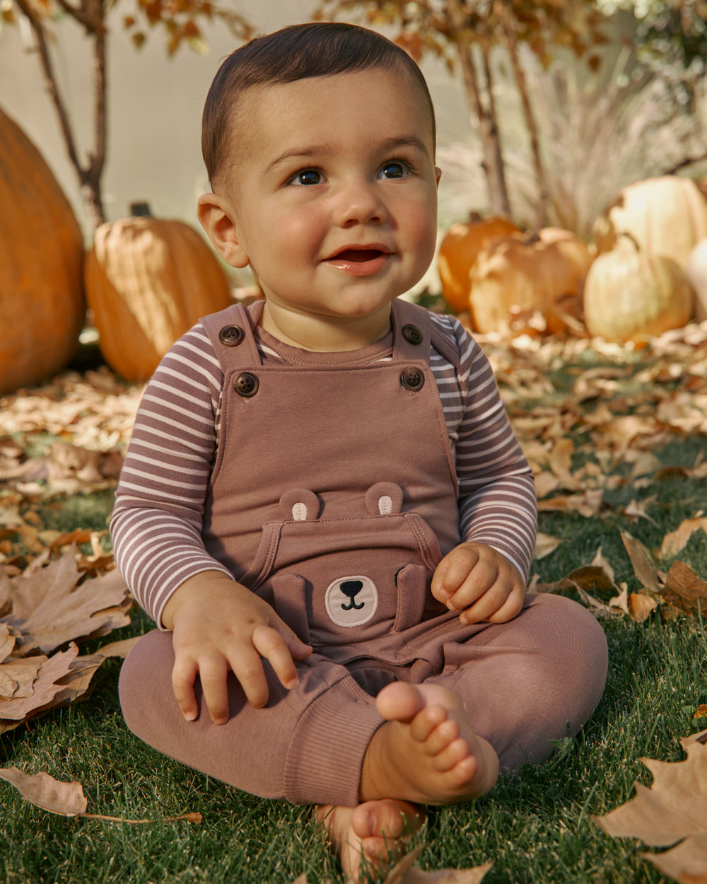 A baby sitting cross-legged on grass outdoors, wearing mauve-brown corduroy overall pants over a striped long-sleeve shirt. Pumpkins and fallen leaves are visible in the background.