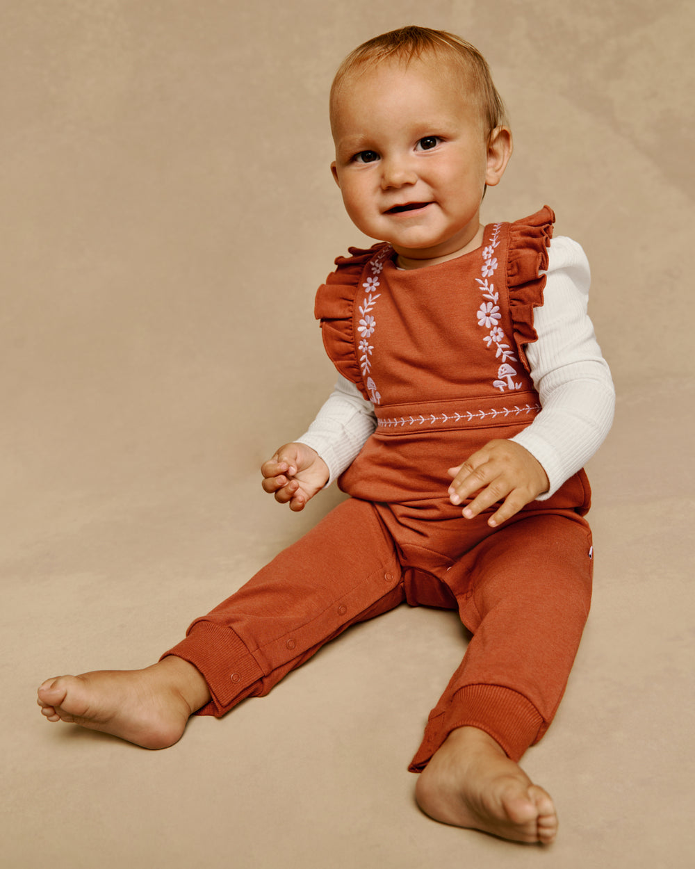 A baby sitting on a beige studio background wearing a rust-orange sleeveless romper over a white long-sleeve shirt. The romper features ruffled shoulders and embroidered floral details along the neckline and chest.