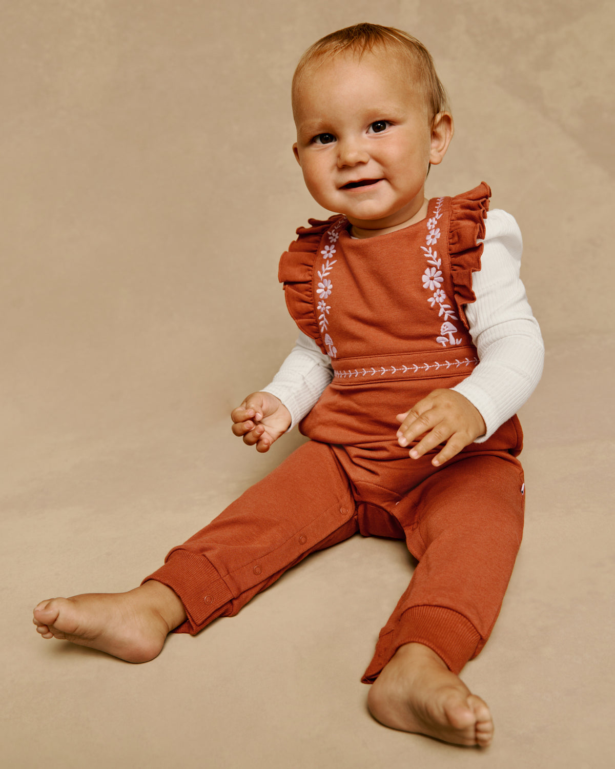 A baby sitting on a beige studio background wearing a rust-orange sleeveless romper over a white long-sleeve shirt. The romper features ruffled shoulders and embroidered floral details along the neckline and chest.
