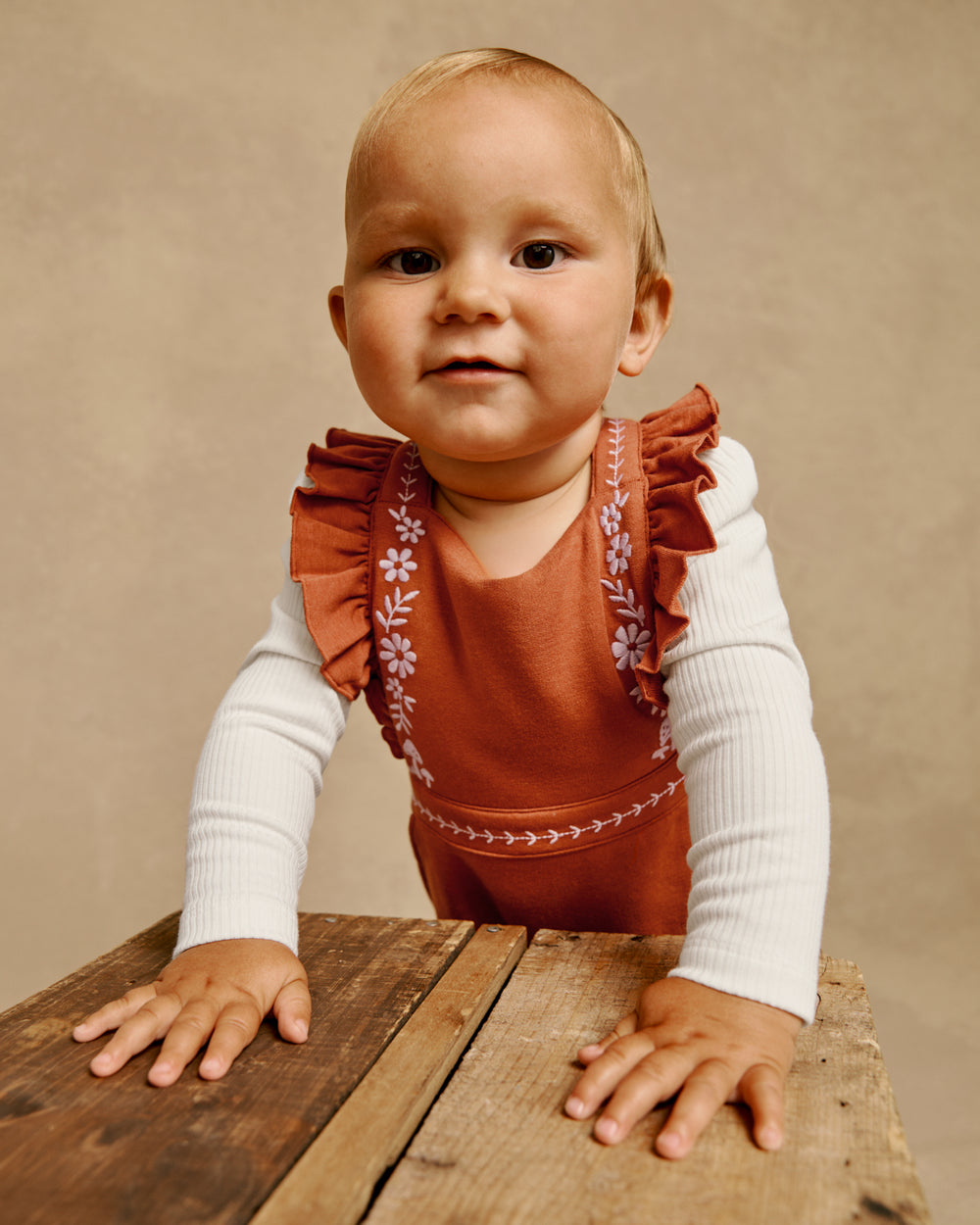 A baby wearing a rust-colored ruffled romper over a white long-sleeve shirt, standing on a rustic wooden table against a warm beige studio backdrop.