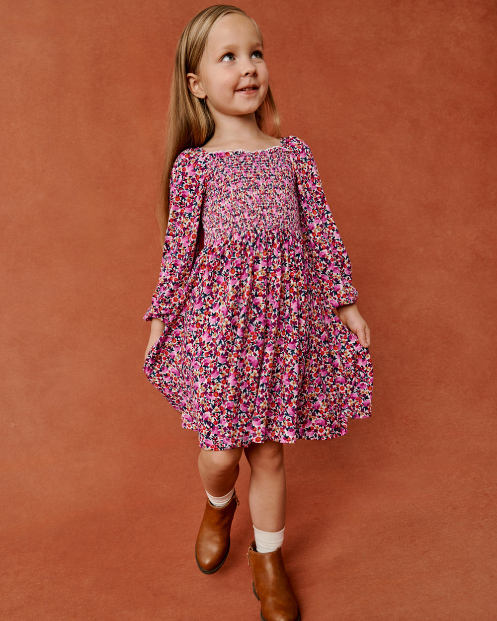 A young girl with long blonde hair wearing a pink and multicolor floral print long-sleeve dress, styled with white socks and brown leather boots, standing against a textured terracotta background.