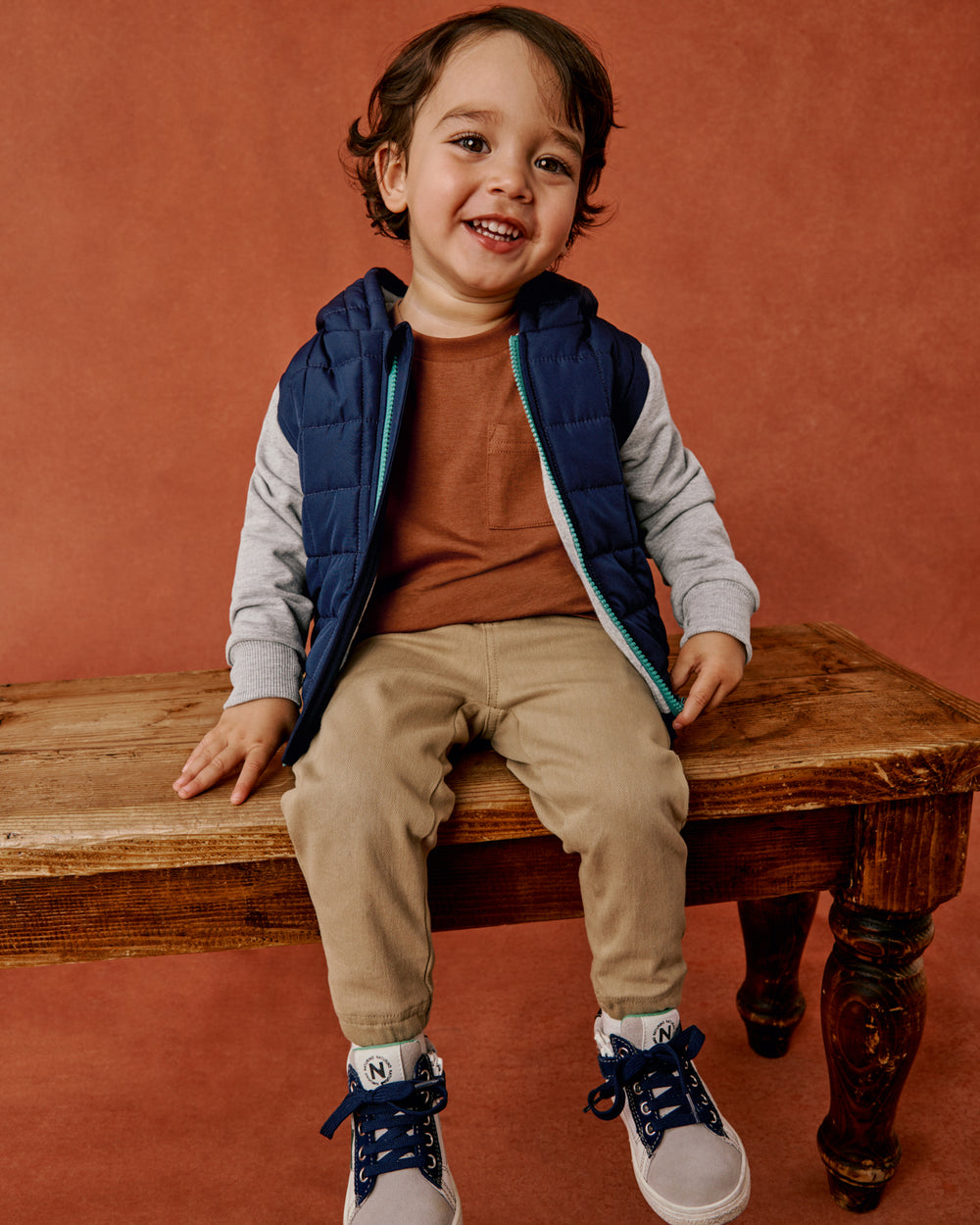 A smiling young child sits on a rustic wooden bench against a warm terracotta background, wearing a navy puffer jacket with gray long-sleeves, tan pants, and navy-and-white sneakers.
