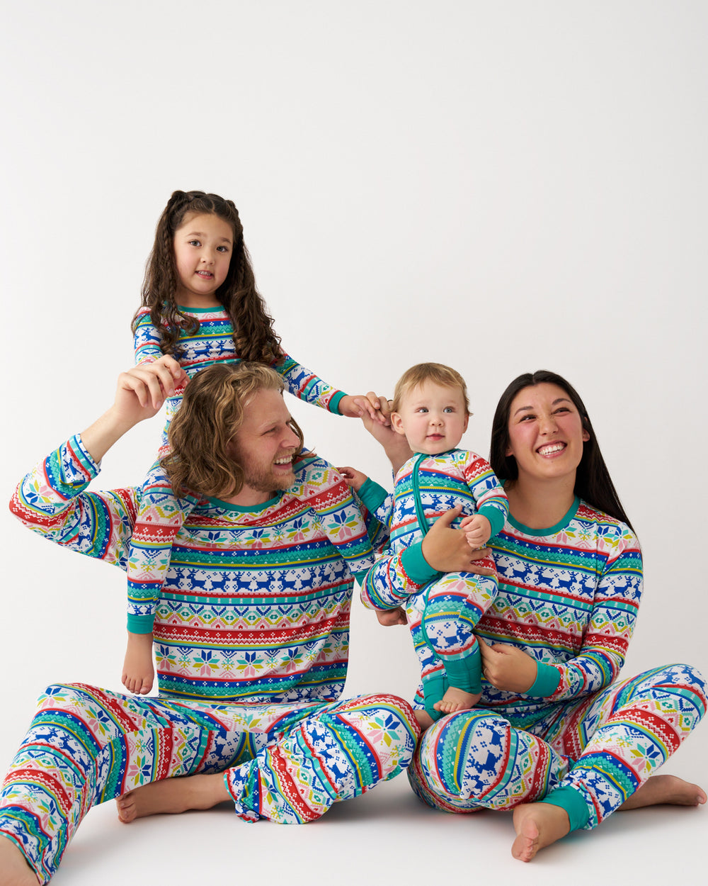 A family of four wearing matching long-sleeve holiday pajamas with a colorful Nordic/Fair Isle pattern, sitting and posing together against a plain white studio background.