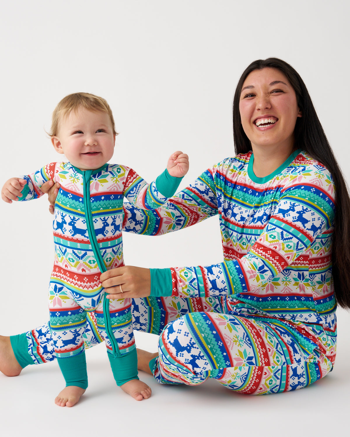 A smiling woman and a baby sitting on a light gray studio background, both wearing matching long-sleeve zip-front pajamas with a colorful geometric pattern in blue, teal, red, yellow, and white.