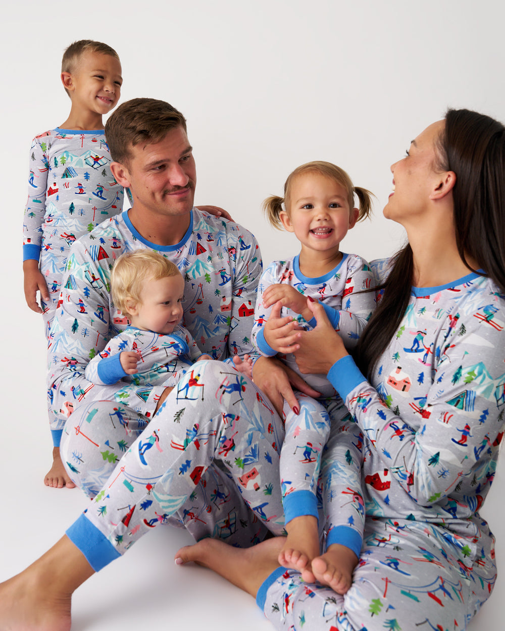 A family of five wearing matching light gray long-sleeve pajamas with a colorful winter sports print, posed together against a plain white studio background.