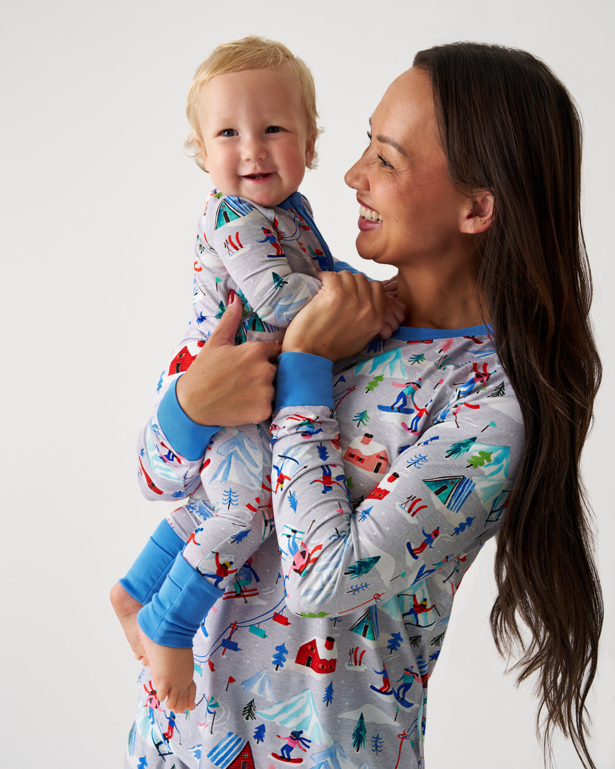 A smiling woman holding a baby wearing matching long-sleeve pajamas with a colorful winter scene print on a light gray background. The pajamas have blue cuffs and collar, photographed against a plain light gray studio background.