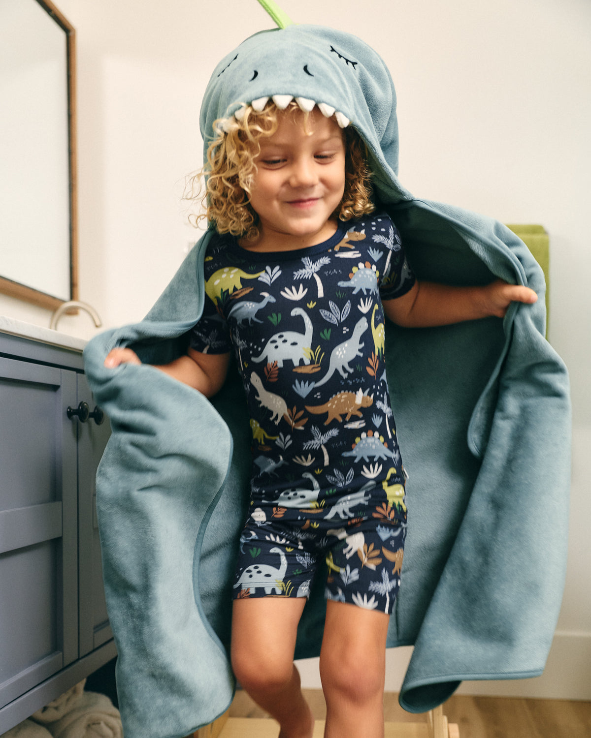 A young child wearing a navy blue dinosaur-print short-sleeve pajama set and a light blue hooded towel with a shark-face hood, standing indoors and holding the towel open.