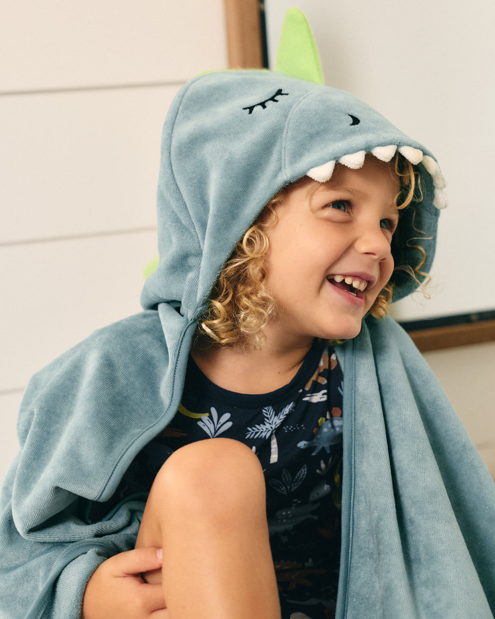 A smiling young child wearing a light blue dinosaur-themed hooded blanket or robe with white teeth details and a green horn, sitting indoors against a light paneled wall.