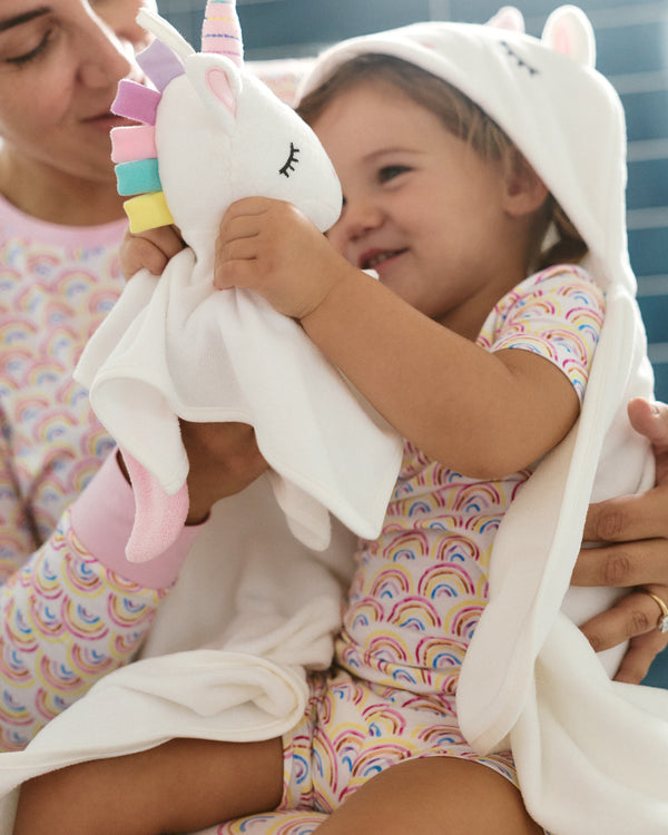 A toddler wrapped in a white hooded towel blanket and wearing a pastel rainbow-patterned pajama set, holding a plush unicorn toy. An adult is partially visible in the background, also wearing matching rainbow-patterned clothing.