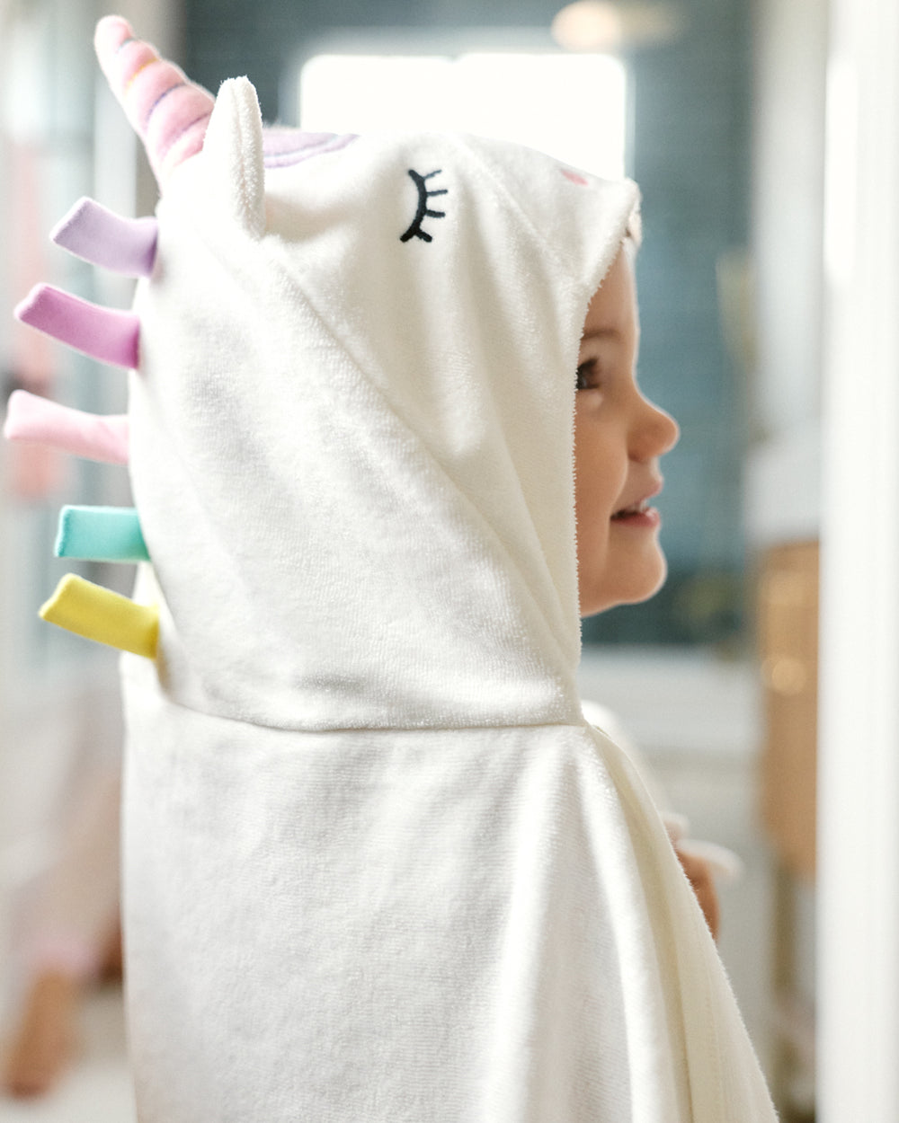 A child wearing a white unicorn-themed hooded bath towel or poncho with pastel horn and tail details, shown in profile against a softly blurred indoor background.