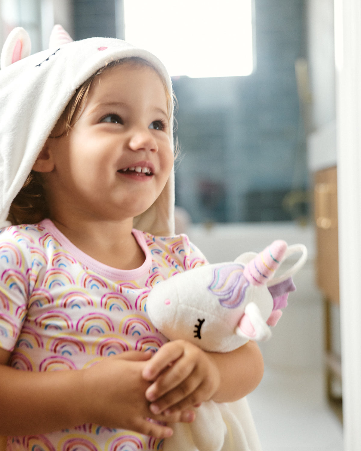 A smiling young child wearing a white hooded towel with bunny ears and a pastel rainbow-patterned shirt, holding a plush unicorn toy in a softly lit indoor setting.