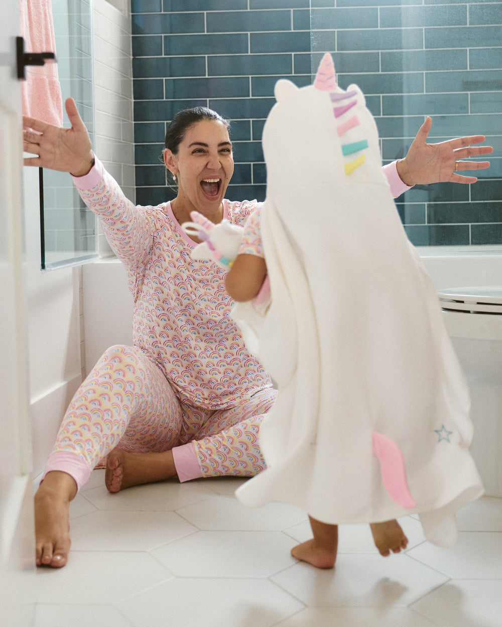 A woman in pink and yellow rainbow-patterned pajamas sits on a white tiled bathroom floor with her arms outstretched, smiling as a child wearing a white unicorn plush blanket walks in front of her.