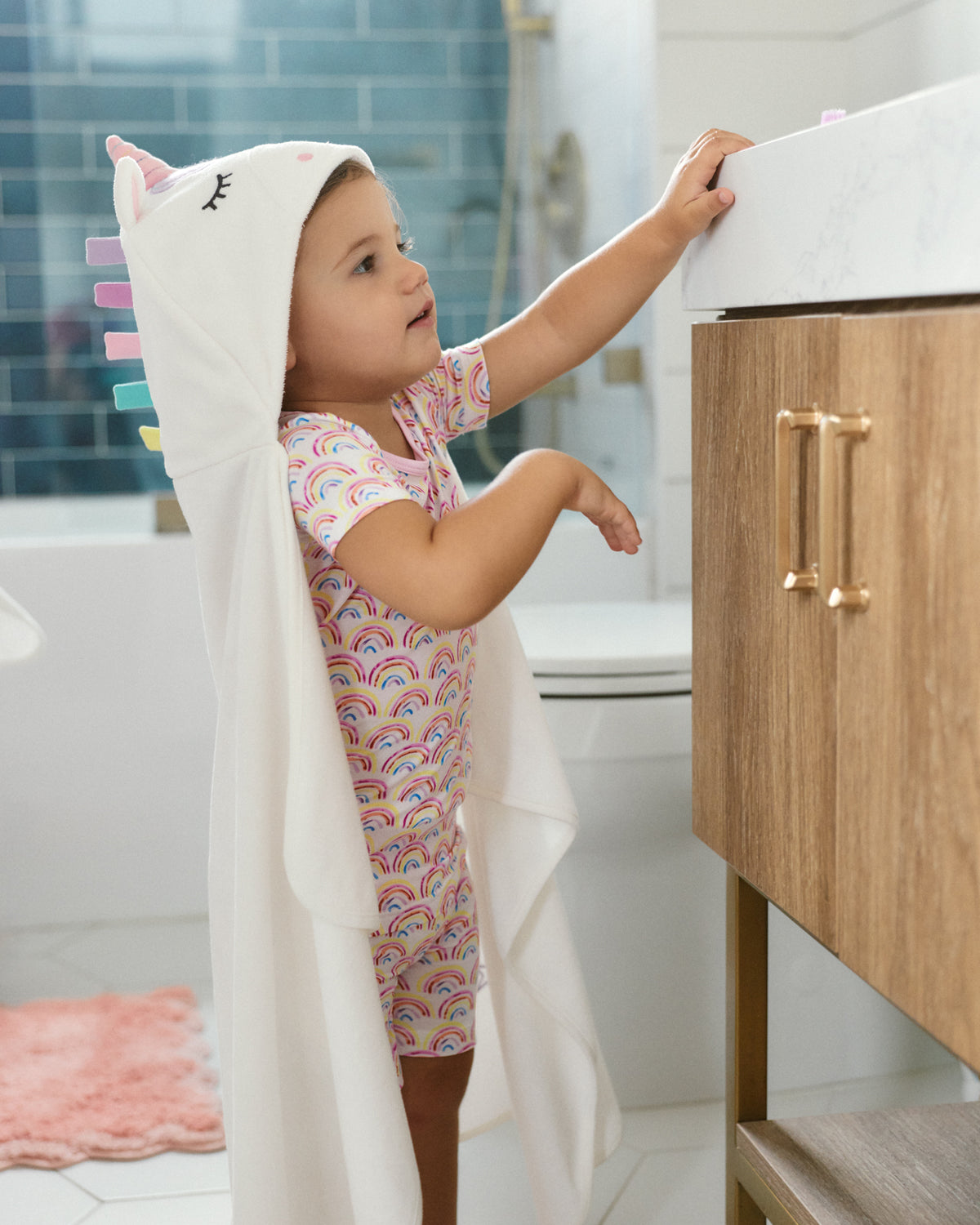 A toddler stands in a bright bathroom wearing a white unicorn-themed hooded towel draped over their body and a pastel rainbow print one-piece swimsuit, reaching toward a marble sink on a wooden vanity.