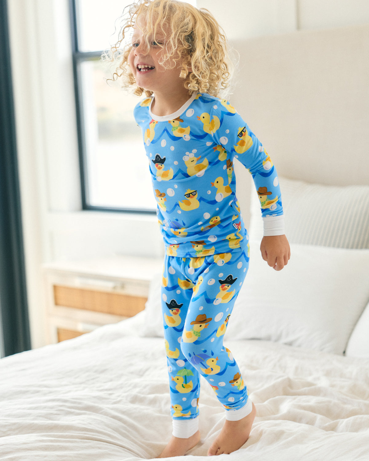 A young child with curly blonde hair stands on a white bed wearing a bright blue long-sleeve pajama set with a yellow rubber duck print. The room has a soft-focus background with a window and neutral-colored headboard.