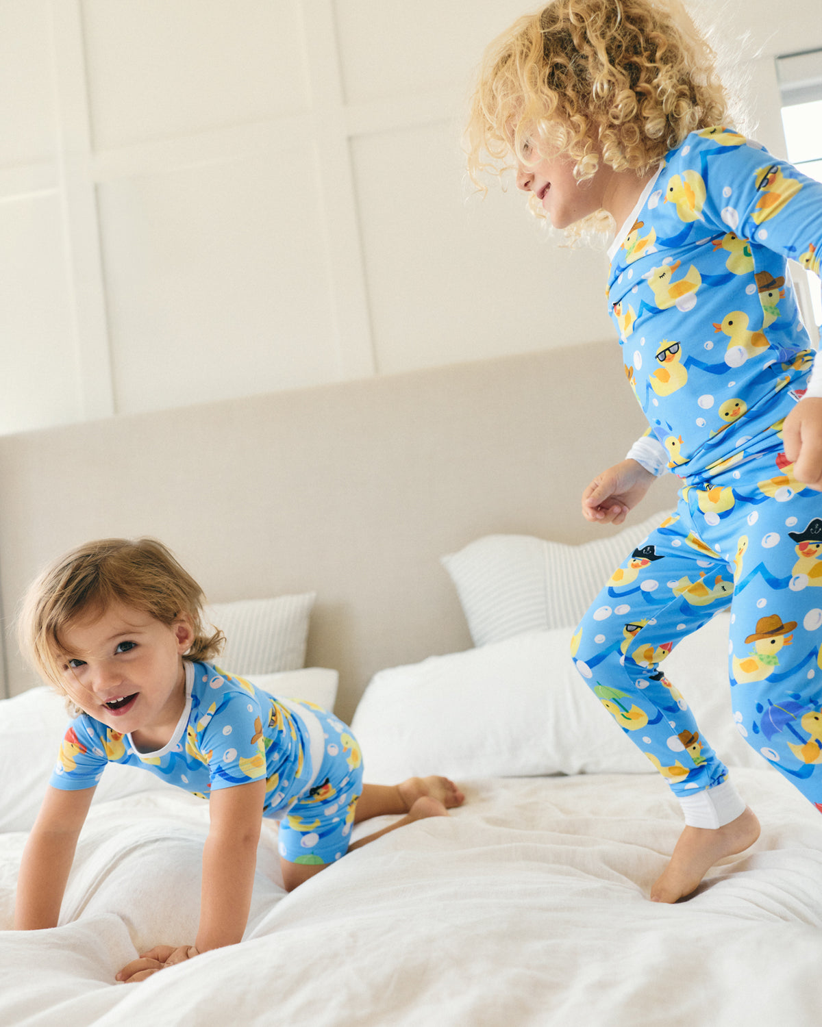 Two children wearing matching blue duck-print pajamas play on a white bed in a bright bedroom.