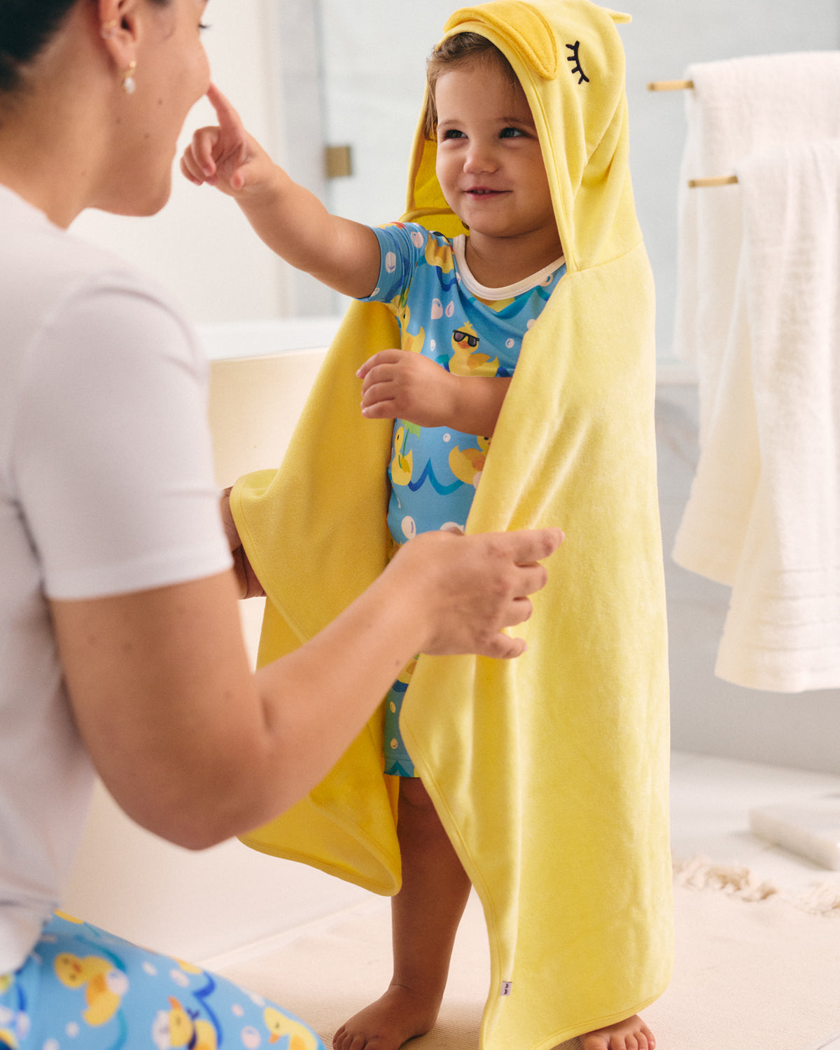 A toddler wearing a blue duck-print onesie stands in a bathroom while wrapped in a bright yellow hooded towel, with an adult kneeling beside them.