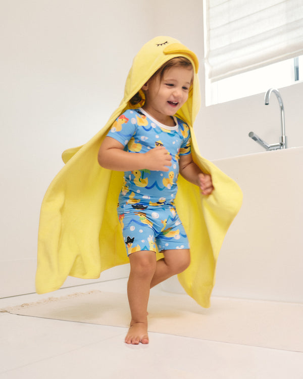 A young child wearing a blue and yellow duck-print pajama set and a large yellow hooded towel draped over their shoulders, standing barefoot in a bright bathroom next to a white bathtub.