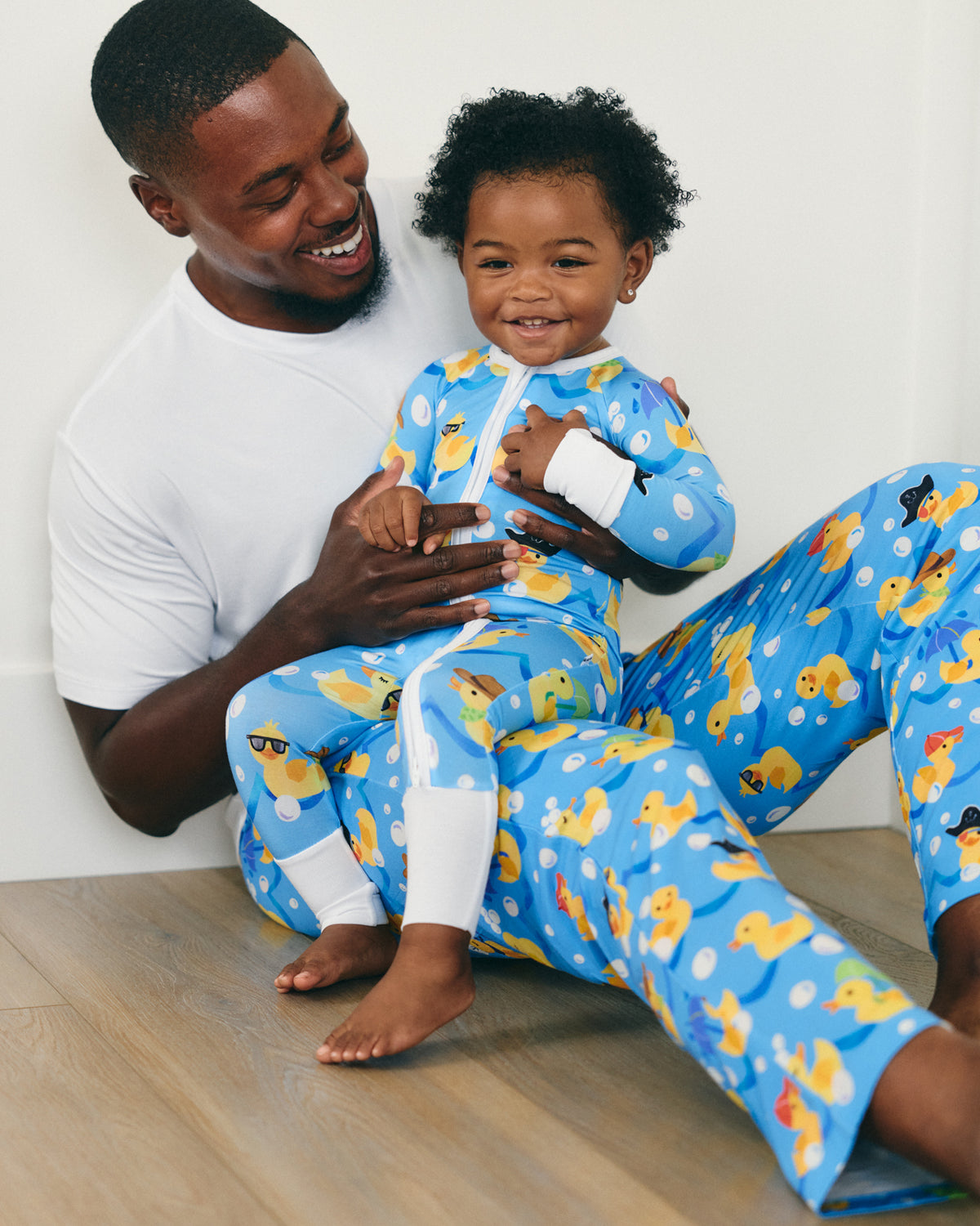 A smiling father and child wearing matching blue duck-print pajamas, sitting together on a light wood floor against a white wall.