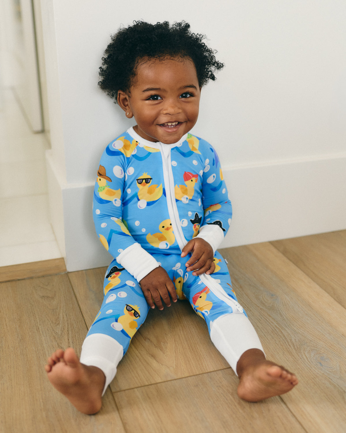 A smiling toddler sitting on a light wood floor against a white wall, wearing a blue zip-front one-piece pajama with a yellow duck print and white cuffs and collar.