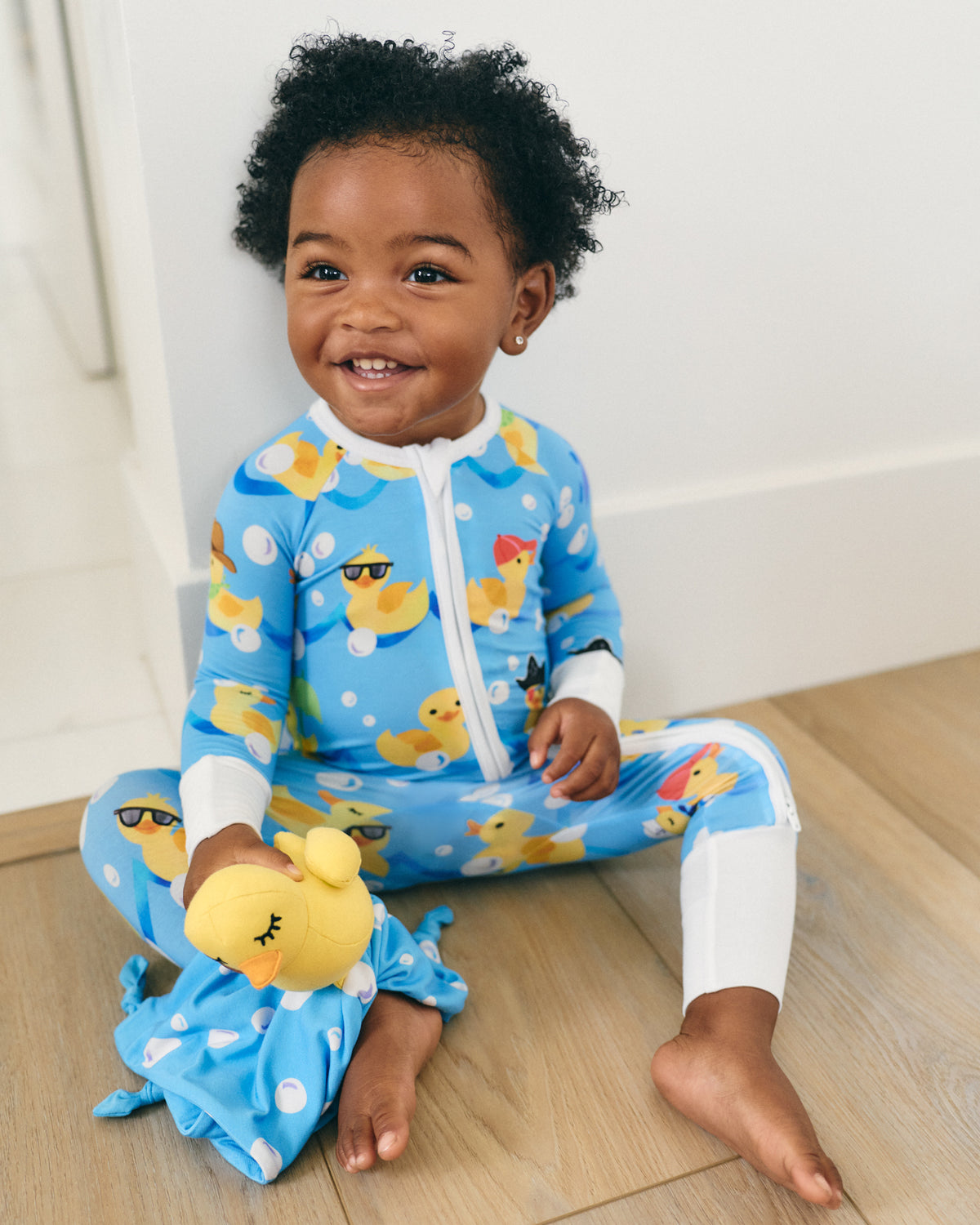 A smiling toddler sits on a light wood floor wearing a blue one-piece pajama with a yellow duck print, white cuffs and collar, and holding a matching blue duck blanket.