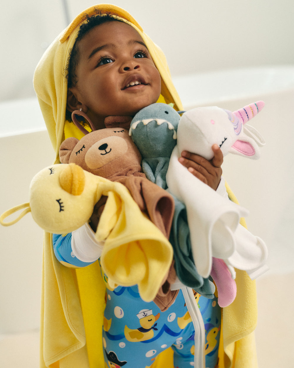 A young child wearing a yellow hooded towel or poncho over blue duck-patterned pajamas, holding a bundle of plush toys including a yellow duck, brown bear, blue shark, and white unicorn.