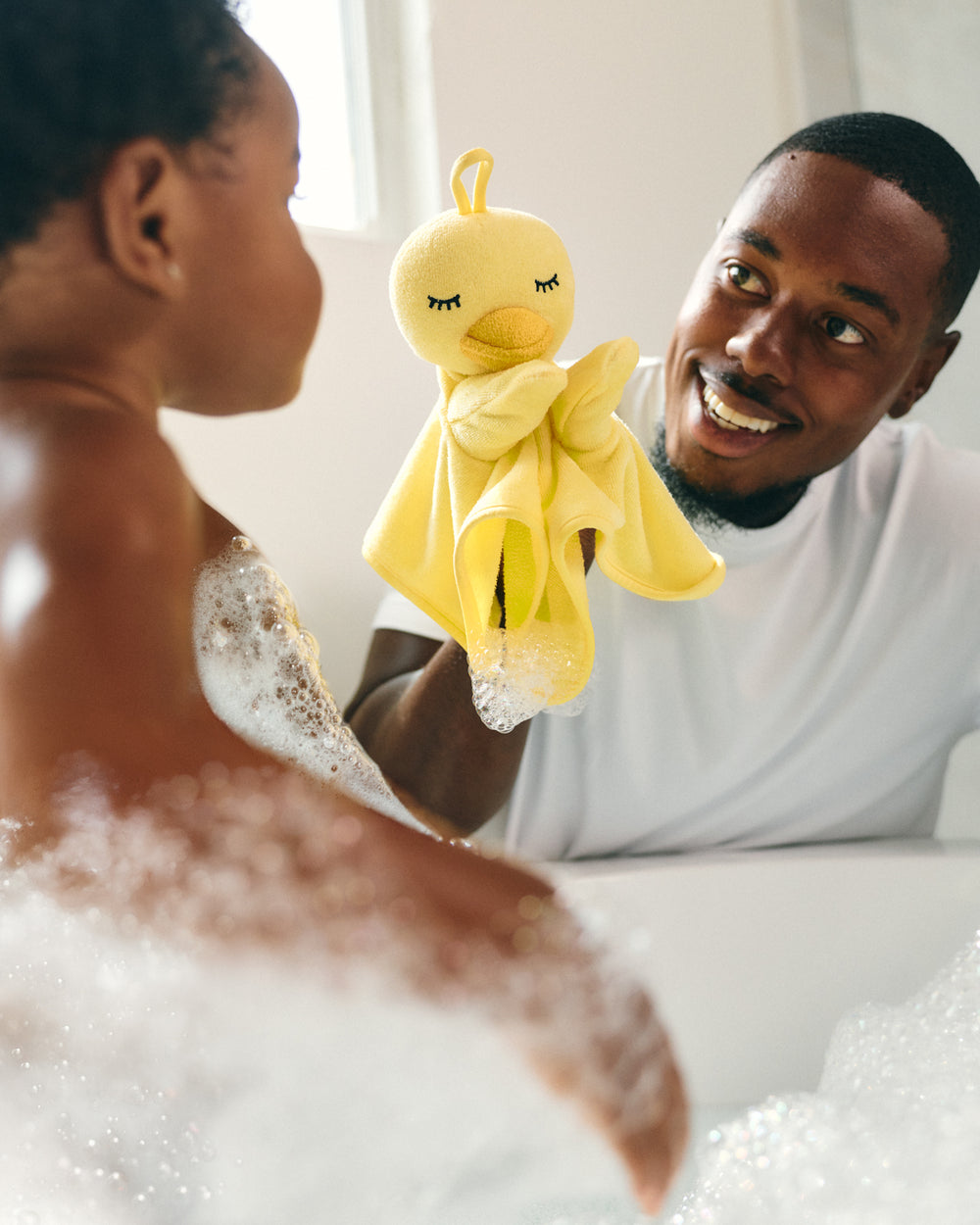 A man holding a yellow plush duck bath toy with a white towel, smiling while a woman sits in a bubble-filled bathtub nearby.