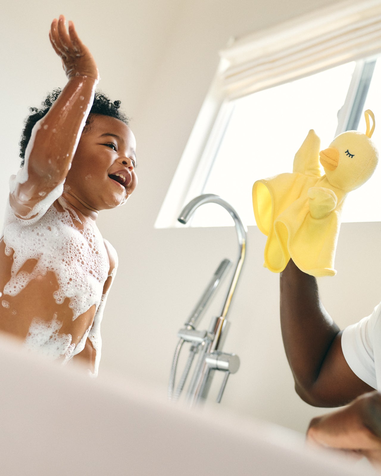 A child taking a soapy bath while playing with a yellow rubber ducky puppet washcloth offered by an adult in a bright bathroom setting.
