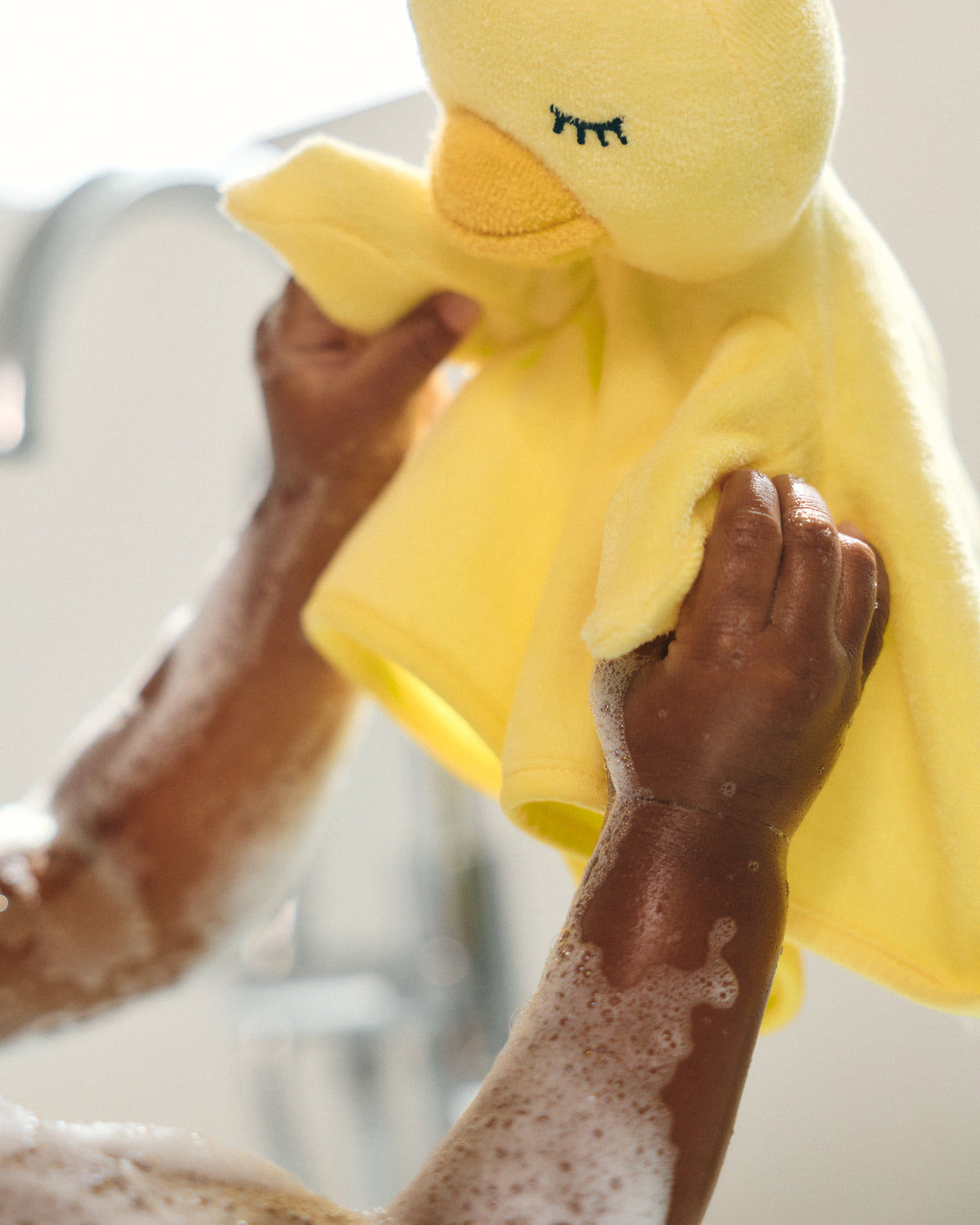 A close-up of a person washing a bright yellow plush duck toy in soapy water, with the person's hands and arms visible against a softly blurred bathroom background.